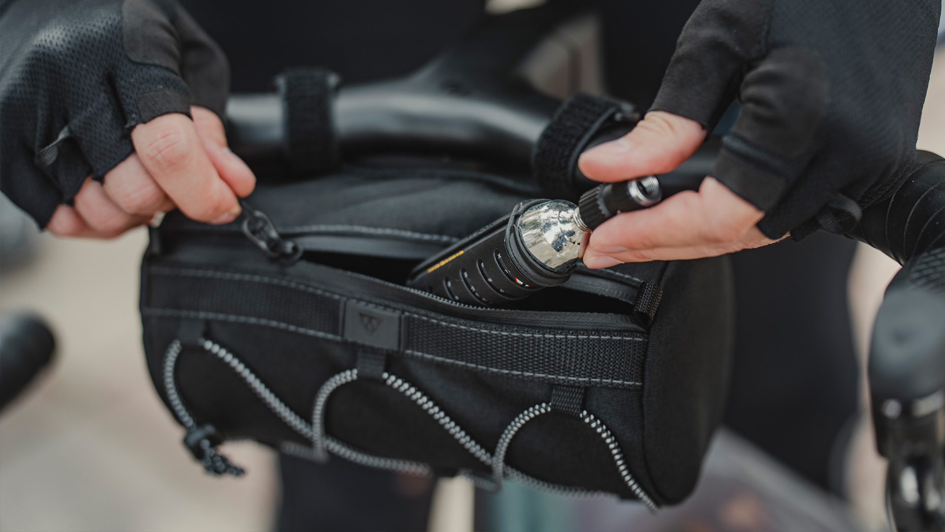 Sacoche de guidon noire ouverte, avec mains gantées rangeant une cartouche de CO2 et un embout de gonflage de vélo