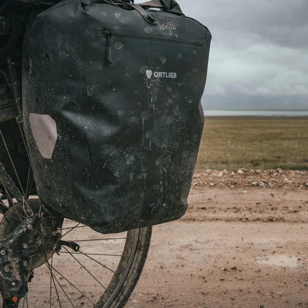 Sacoche vélo Ortlieb Back-Roller Zip-Pocket noire, très sale, fixée sur la roue arrière sur un chemin boueux en pleine campagne.