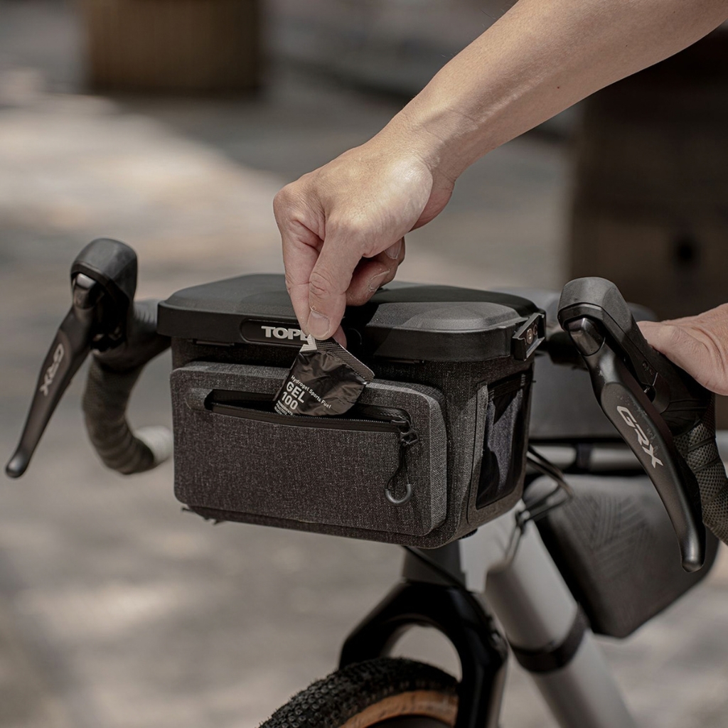 Sacoche de cintre Topeak Elementa Dry BarBag vélo noire avec poche zippée, fixée entre les cocottes, une main y glisse un gel énergétique.
