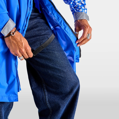 Giacca antipioggia blu aperta indossata sopra un paio di jeans, con le mani che regolano il capo e sfondo bianco neutro