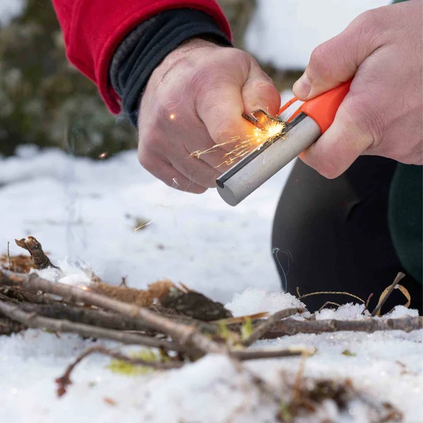 Gros plan sur des mains allumant feu avec un pierre à feu Mag Stricker SOL, étincelles sur fond de neige et brindilles.